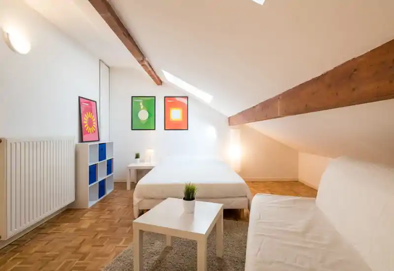 Chambre mansardée lumineuse et accueillante avec poutres apparentes en bois, lit double en linge blanc, éclairage doux par lampe et fenêtre de toit, et mobilier minimaliste.