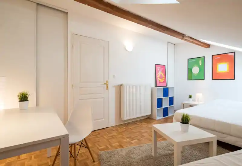 Chambre mansardée lumineuse et moderne avec grand lit blanc, mobilier minimaliste, parquet en bois et affiches décoratives — idéale pour un séjour confortable.