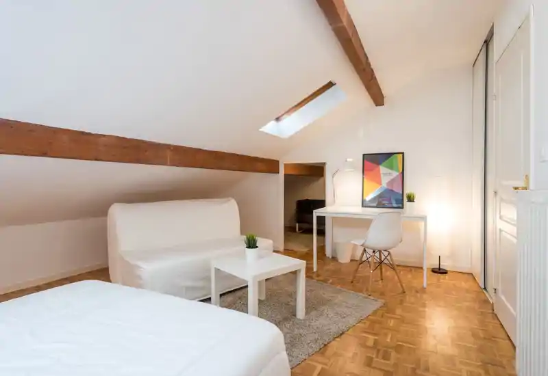 Chambre mansardée lumineuse avec lit au premier plan, petit canapé blanc et bureau minimal sous une fenêtre de toit ; poutres en bois et décor épuré pour une ambiance chaleureuse.