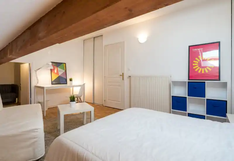 Chambre lumineuse et aérée avec grand lit blanc, poutre en bois apparente et un petit espace bureau. Décor moderne et épuré avec rangements bleus et éclairage doux, accueillant pour se reposer ou travailler.