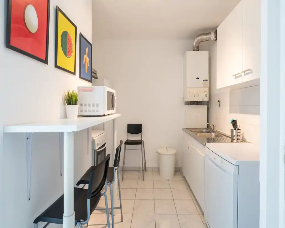 Compact, modern kitchenette with white cabinets, a double sink, dishwasher, microwave and a small breakfast bar with stools — clean and practical for short stays.