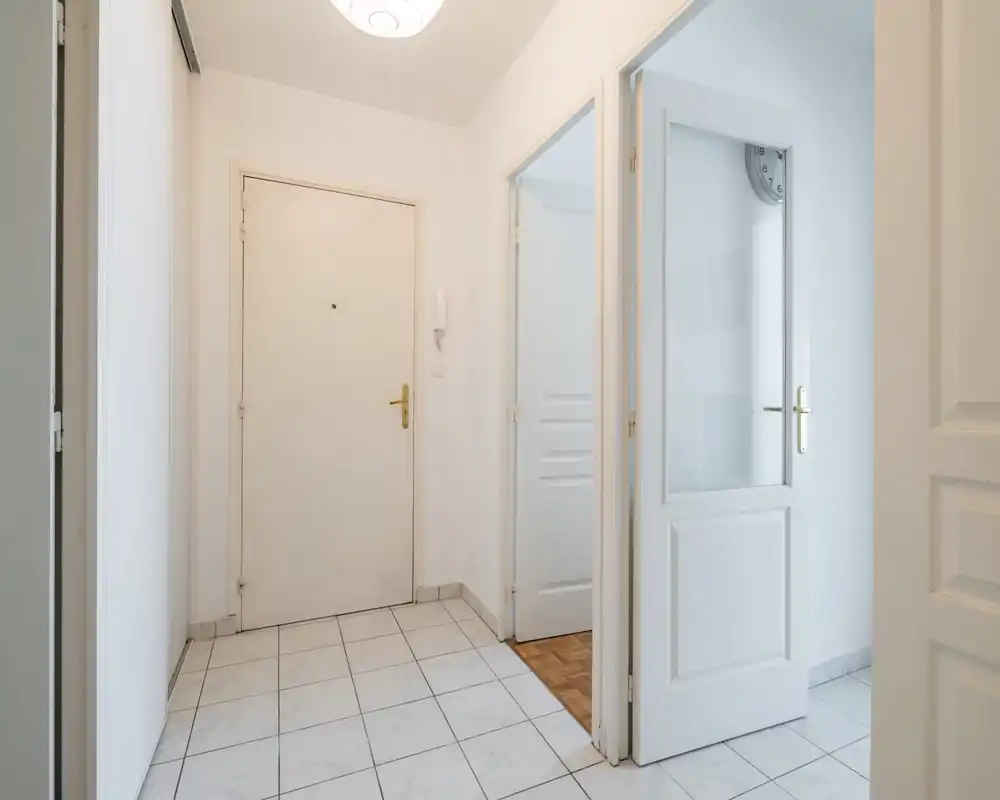 Bright, clean entryway with tiled floor, white walls and a solid front door with brass handle; multiple interior doors lead to adjacent rooms.