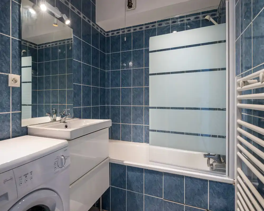 Clean, well-lit bathroom with blue tiled walls, a bathtub with frosted glass screen, sink with vanity and a washing machine visible beside it.