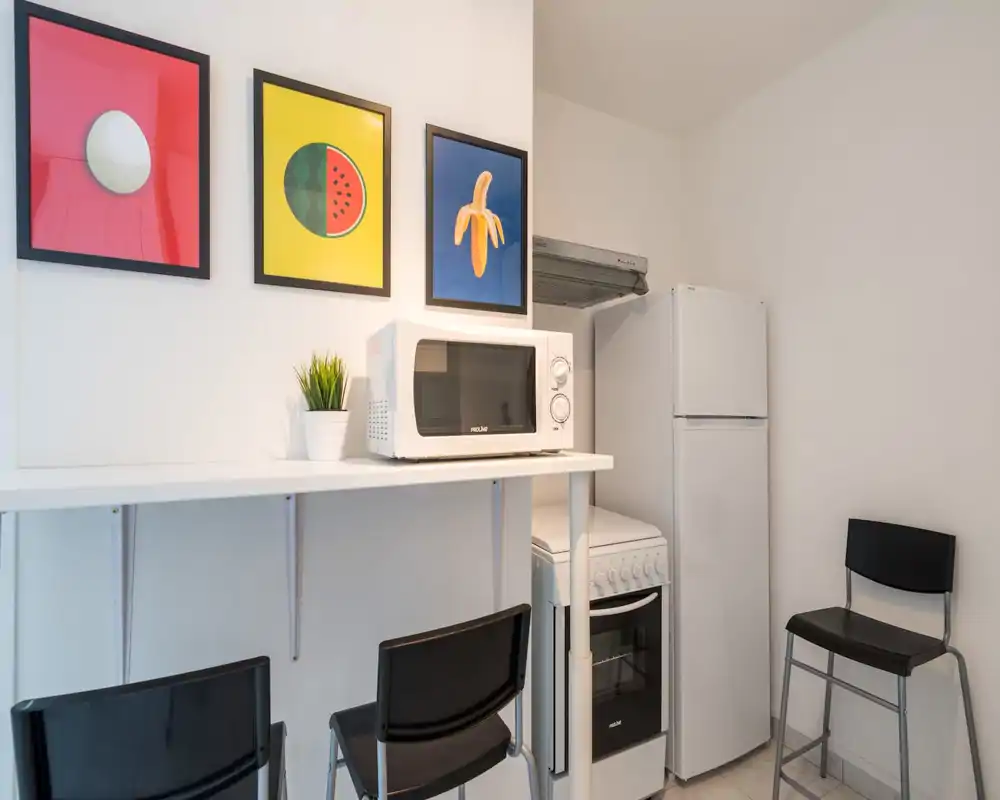 Compact kitchenette with a microwave, stove and refrigerator, accented by colorful framed prints and a small plant; bright, clean and efficiently arranged for short stays.