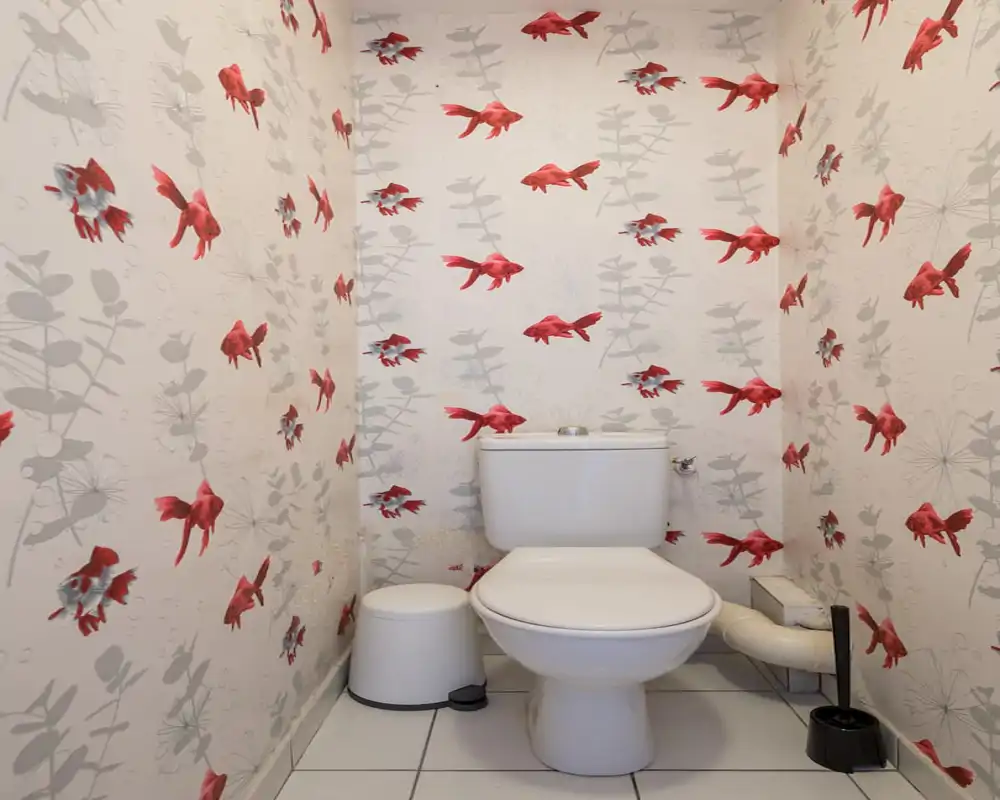 Compact bathroom with a white toilet, matching trash bin and brush, decorated with playful red-goldfish patterned wallpaper and white tiled floor.