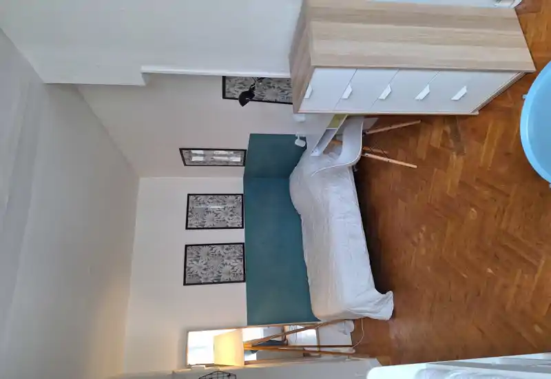 Sunny, well-styled bedroom with a single bed against a teal-accented headboard, small desk and chair, framed wall art and warm wooden parquet flooring.