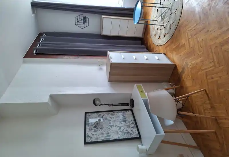 Bright, minimal room corner with a white desk, chair, tall drawers and soft curtains; wooden parquet and a round woven rug add warmth.
