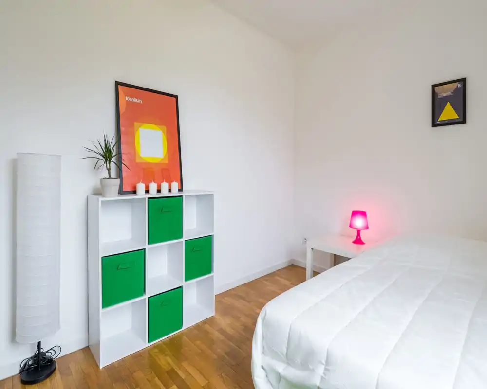 Chambre lumineuse et minimaliste avec une couette blanche au premier plan, une étagère cubique avec tiroirs verts, des affiches modernes et une lampe de chevet rose apportant une touche de couleur.
