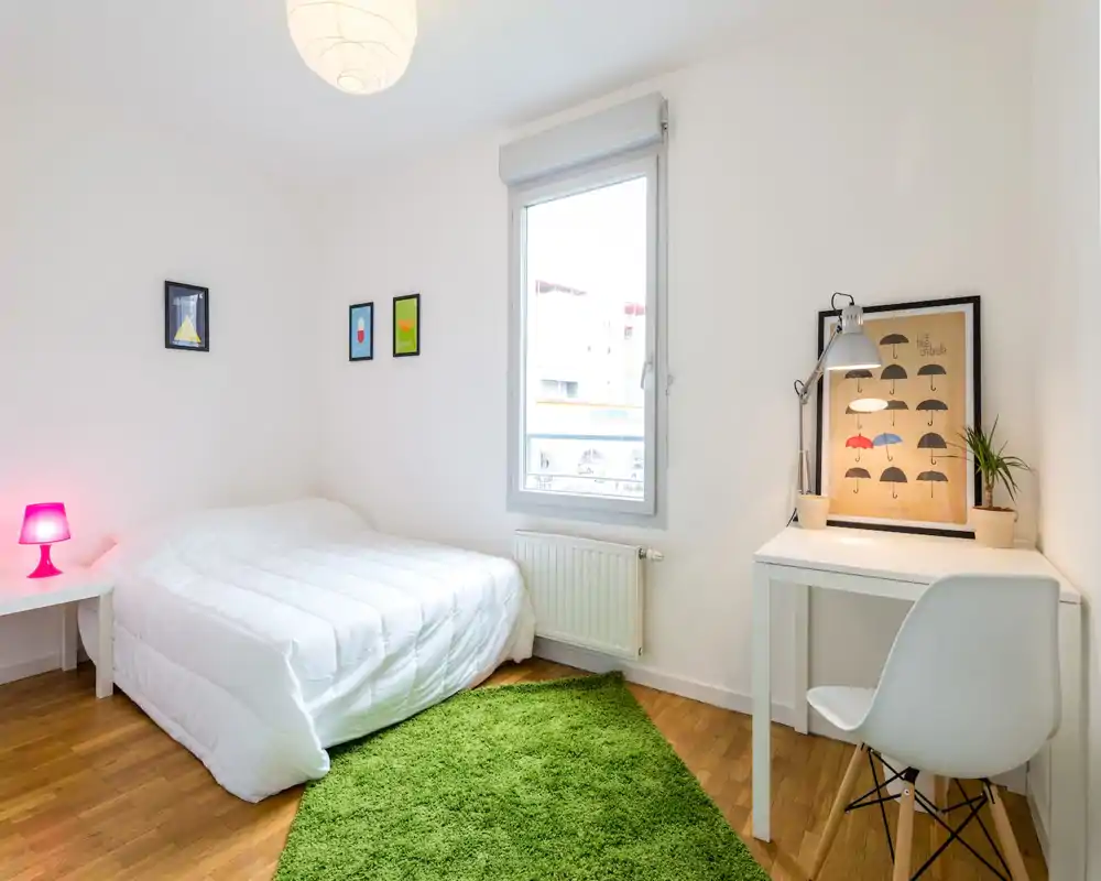 Chambre lumineuse et minimaliste avec un lit double confortable, un tapis vert comme accent et un petit bureau près de la fenêtre apportant de la lumière naturelle.