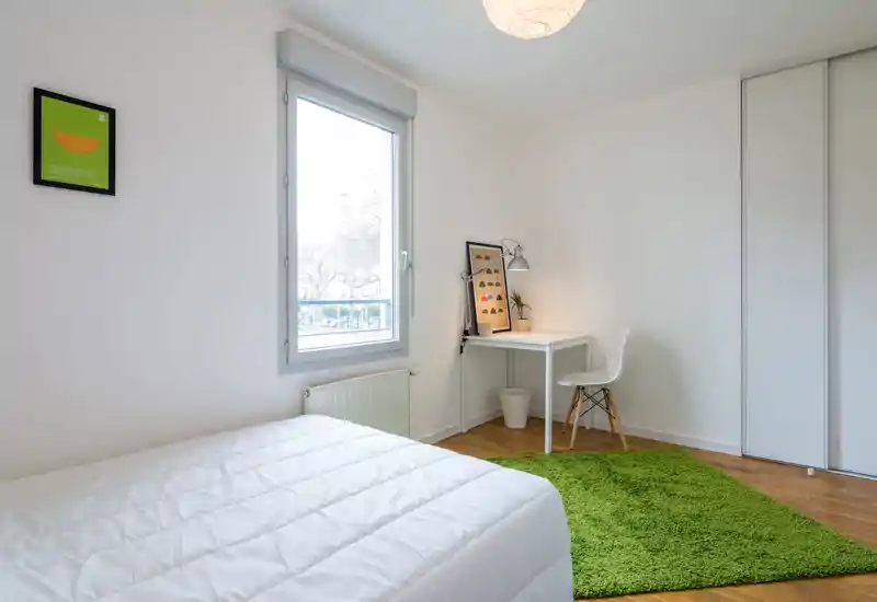Bright, minimalist bedroom with a white bed, a small desk and chair by the window, and a vivid green rug adding color.