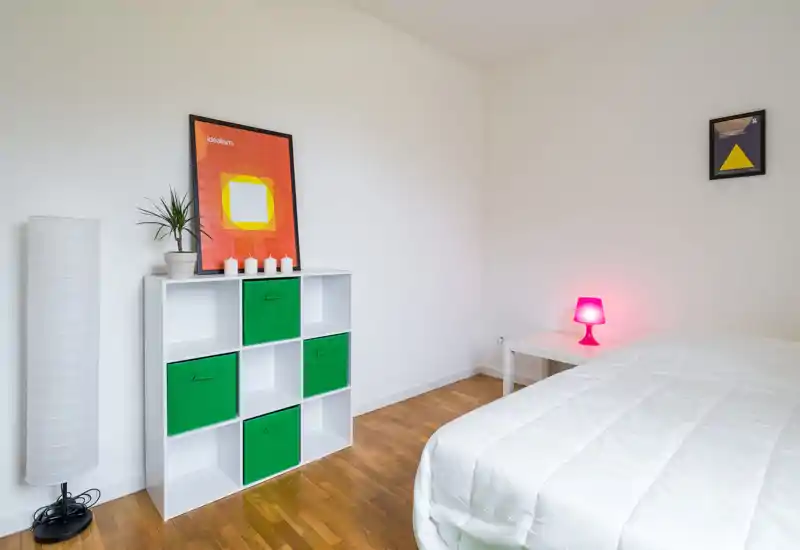 Minimal, bright bedroom with a white quilted bed in the foreground, a cubic storage unit with green fabric drawers, modern artwork and a pink bedside lamp adding color accents.