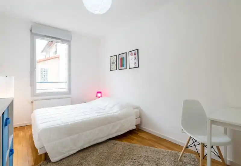 Chambre lumineuse et minimaliste avec un lit double bien fait, un tapis moelleux et une fenêtre apportant une belle lumière naturelle. Décoration moderne et épurée avec cadres et petite lampe de chevet, idéale pour les locations courte durée.