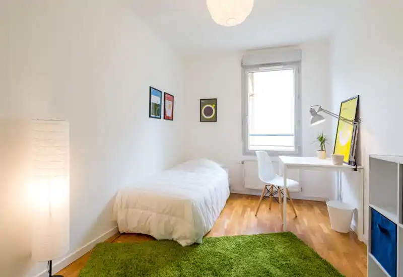Bright, minimalist single bedroom with a neatly made bed, a study desk by the window and warm, natural light. Clean white walls, a green rug and simple framed prints create a fresh, modern feel.
