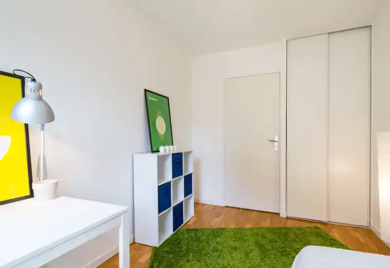 Minimalist bedroom corner with a white desk, lamp and storage cube unit. A green rug and simple decor add a fresh, cozy touch; the bed is partially visible in the foreground.