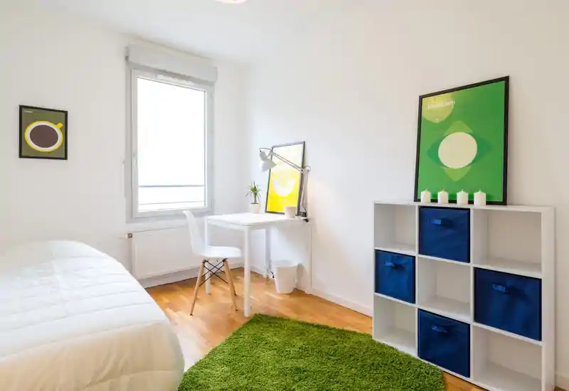 Bright, minimalist bedroom with a neatly made bed, a small white desk by the window, and a shelving unit with blue storage bins. Natural light and a green rug add a fresh, welcoming feel.