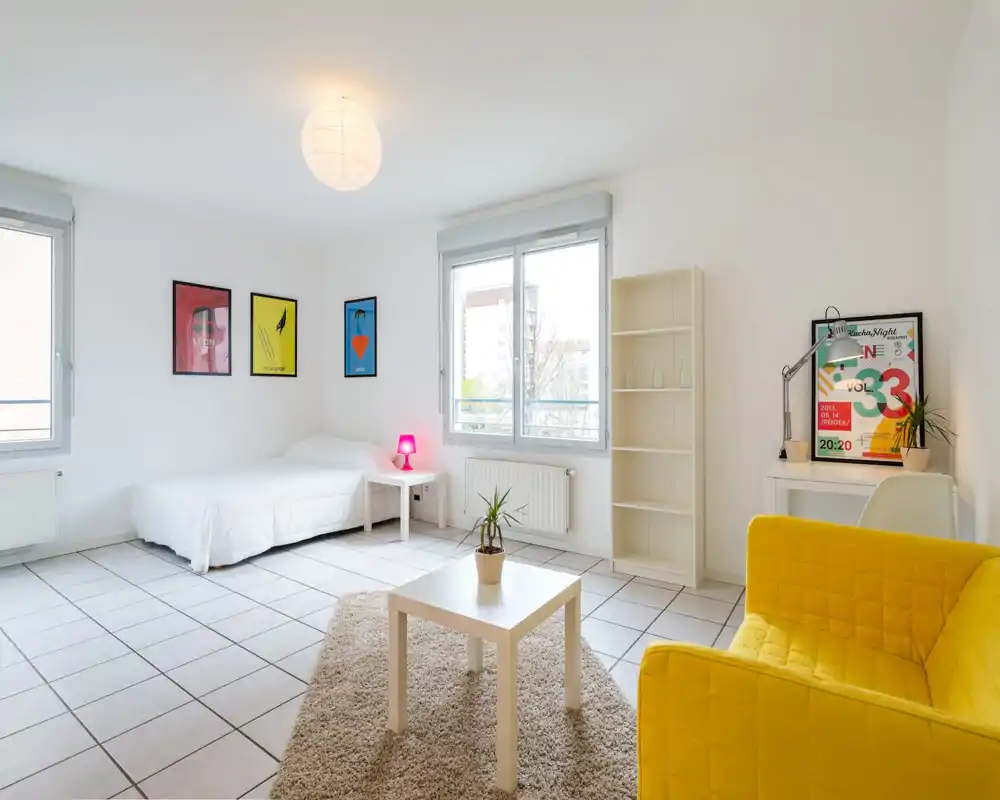 Bright, modern bedroom-studio with a comfortable white bed, large windows letting in abundant natural light, colorful wall art and a cozy rug — ideal for short-term rentals.