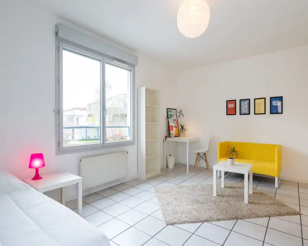 Bright, modern bedroom/studio with a large window, white furnishings, a cozy rug and a cheerful yellow sofa. Ideal for listings emphasizing natural light and minimalist decor.
