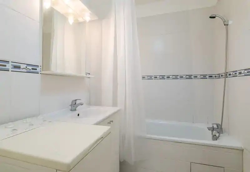 Salle de bain lumineuse et propre avec baignoire, douche murale et lavabo avec miroir ; carreaux blancs neutres pour une atmosphère fraîche et minimaliste.