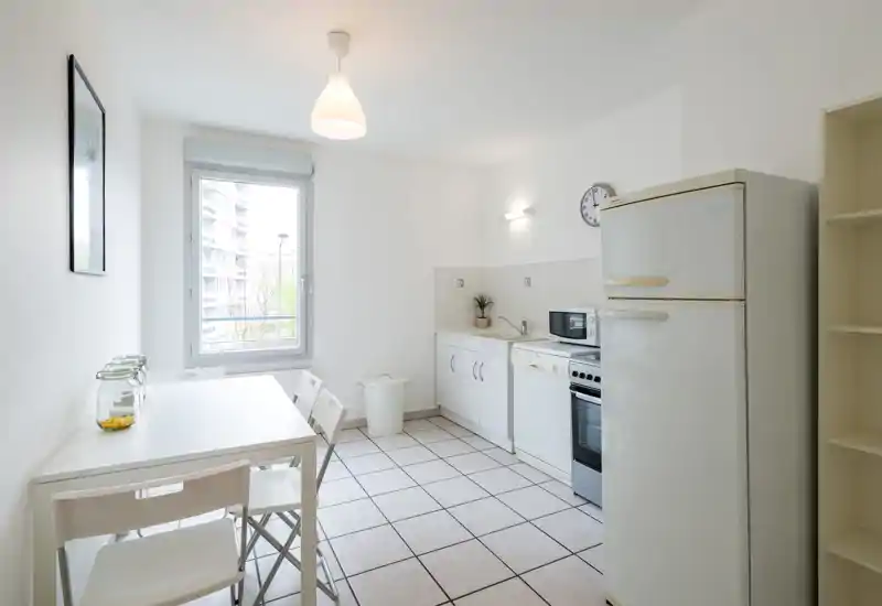 Bright, minimalist kitchen with a dining table for four, white cabinets, a freestanding fridge, and natural light from a large window — ideal for a compact apartment.
