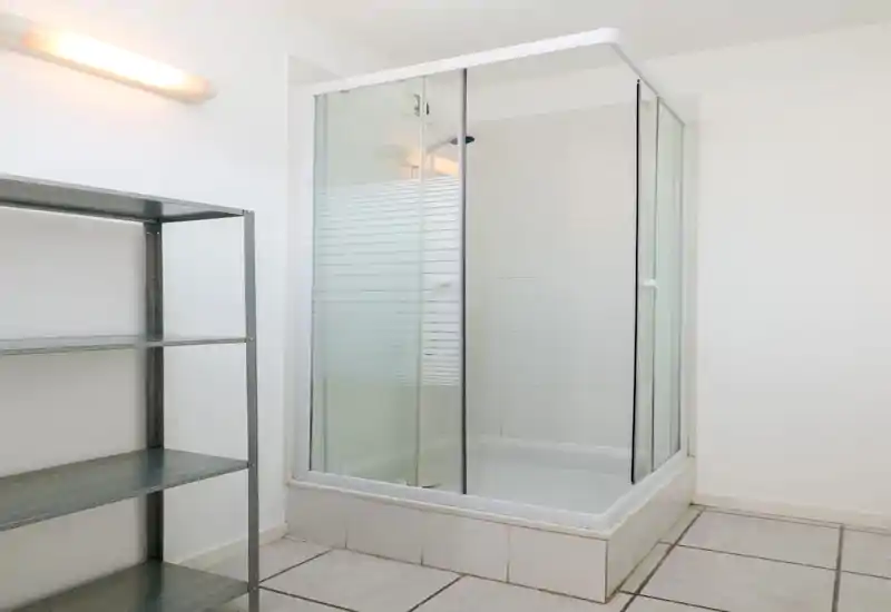 Compact bathroom with a glass-enclosed shower cabin, white tiles and simple metal shelving — bright and functional.