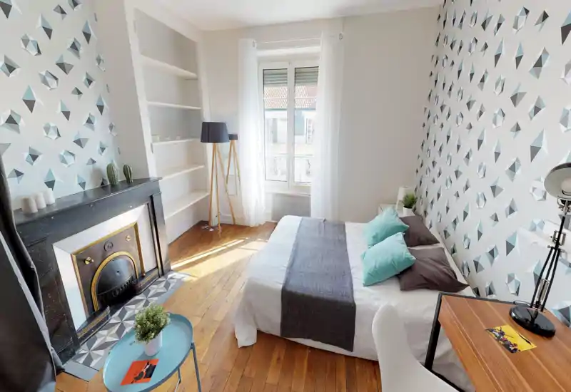 Chambre lumineuse et stylée avec papier peint à motifs, lit double bien fait avec coussins turquoise, parquet et une cheminée décorative — idéale pour une location cosy.