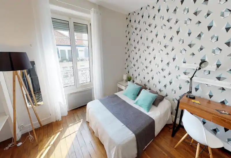 Chambre lumineuse et stylée avec lit double, mur à motif, lumière naturelle depuis une grande fenêtre et un petit bureau — parfaite pour locations courte durée ou un séjour confortable.