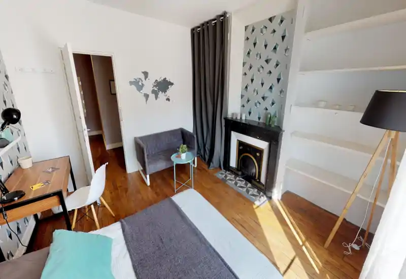 Chambre lumineuse et élégante avec lit bien visible au premier plan, un petit canapé et une cheminée décorative. Parquet, papier peint à motifs et lumière naturelle offrent une ambiance chaleureuse et moderne.