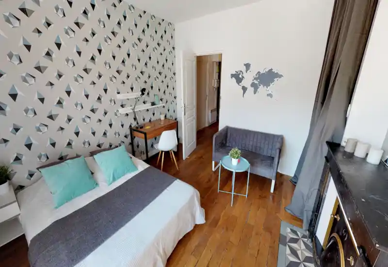 Chambre lumineuse et stylée avec mur à motifs, lit double avec coussins turquoise, petit bureau et coin salon compact — parquet chaud et décoration soignée pour une ambiance accueillante.