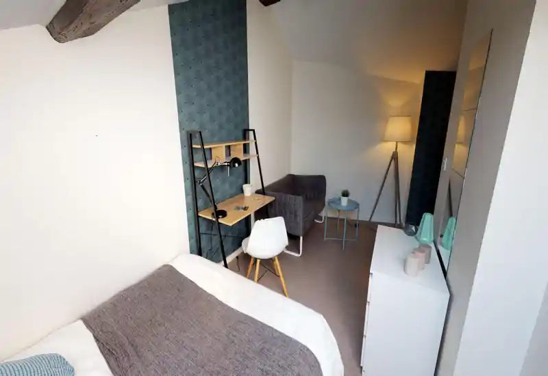 Bright, styled bedroom with a visible double bed in the foreground, a compact desk and chair against a patterned accent wall, and a small sofa with side table and floor lamp — a cozy, modern space ideal for short stays.