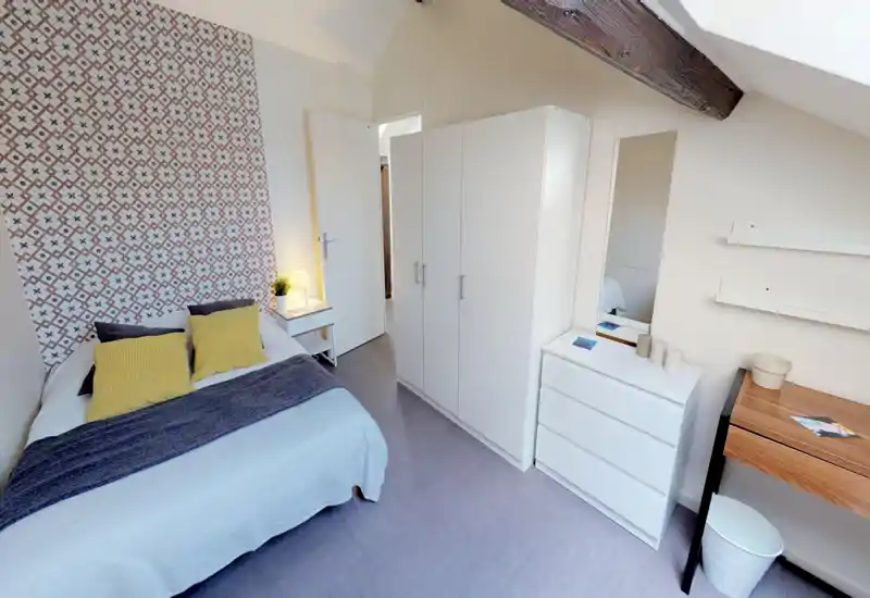 Camera da letto luminosa e ben arredata con letto matrimoniale, testiera decorata, comodino con lampada e armadio bianco. La stanza ha una trave in legno e luce naturale da una finestra a tetto, creando un’atmosfera accogliente.