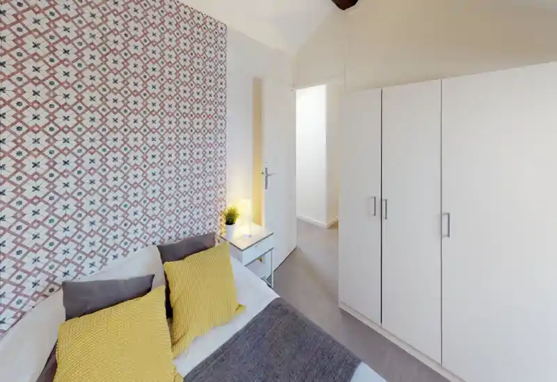 Camera da letto luminosa e ben allestita con una parete decorativa, tessili accoglienti e un armadio compatto. Il letto è ben visibile con cuscini decorativi e una lampada da comodino che crea un'atmosfera calda.