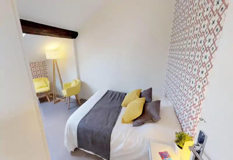 Camera da letto luminosa e accogliente con letto matrimoniale in lino bianco, runner grigio e cuscini gialli; parete decorativa, lampada d’angolo e due poltroncine gialle per un ambiente curato.