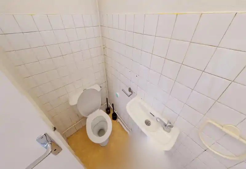 Compact tiled bathroom with a toilet and a small wall-mounted sink, neutral white tiles and a simple layout—suitable for a single-occupant flat.