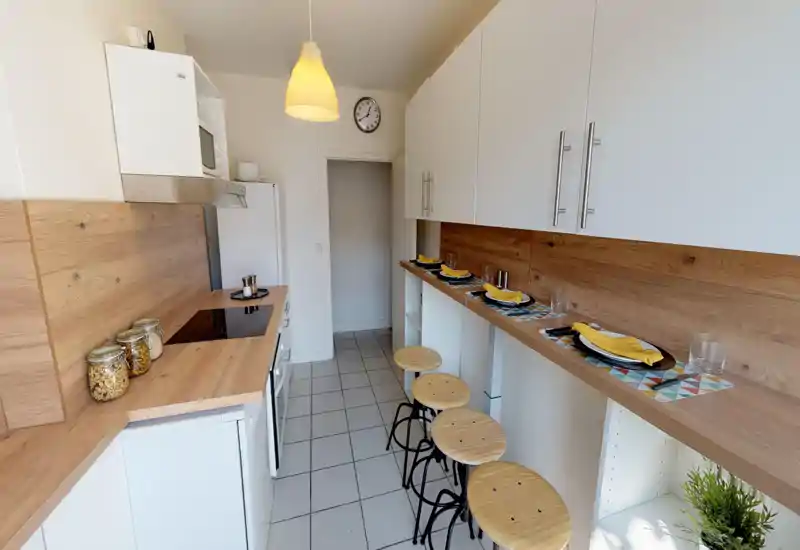 Cucina moderna, luminosa e stretta con piano e paraschizzi effetto legno, bancone con sgabelli e tovagliette apparecchiate — spazio pratico e accogliente per pasti veloci.