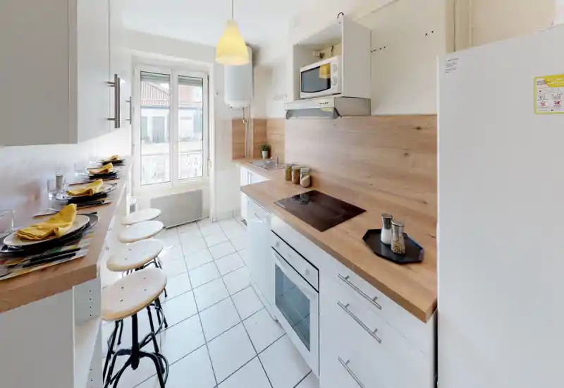 Cuisine en longueur lumineuse et moderne avec plans de travail en bois, plaque à induction et bar avec tabourets — idéale pour un logement urbain compact.