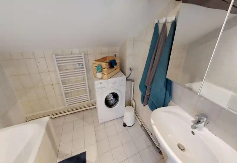 Compact tiled bathroom with a bathtub, sink, wall-mounted towel radiator and a washing machine; neutral tiles and teal towels give a clean, functional look.