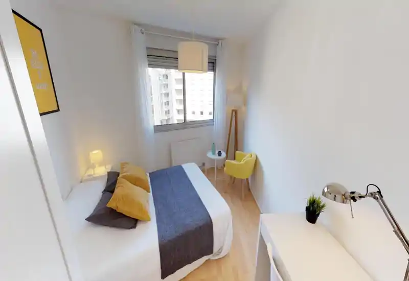 Bright, modern bedroom with a double bed dressed in white linens, decorative cushions and a grey runner; window provides natural light and a cozy yellow accent chair sits by a small side table.
