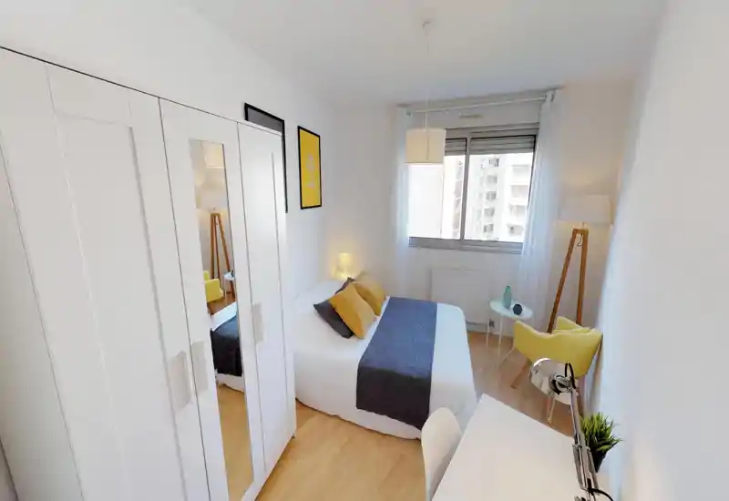 Bright, modern bedroom with a double bed dressed in white linens and a gray runner, warm mustard cushions, built-in wardrobe with mirrored door, desk workspace and a cozy reading corner with a yellow armchair and floor lamp.