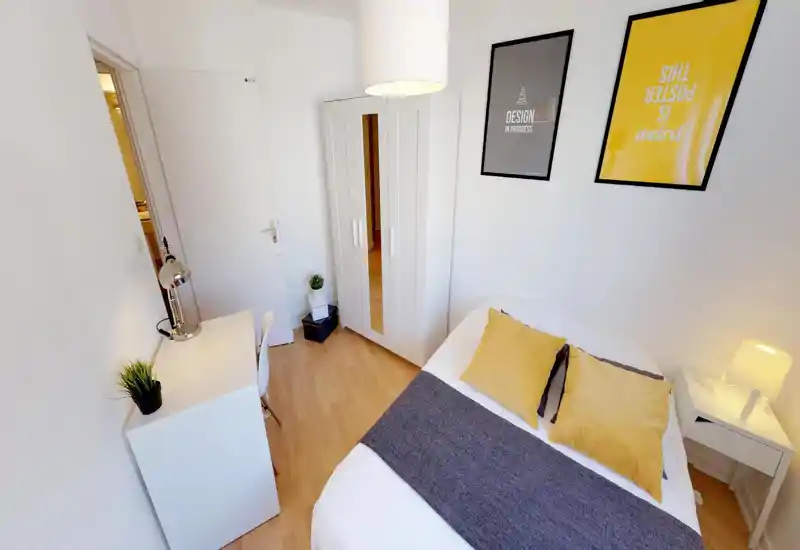 Bright, well-styled bedroom with a double bed dressed in white linens, yellow accent cushions and a gray throw. The room includes a white wardrobe, small bedside table with lamp and a compact desk — ideal for a cozy single or couple.