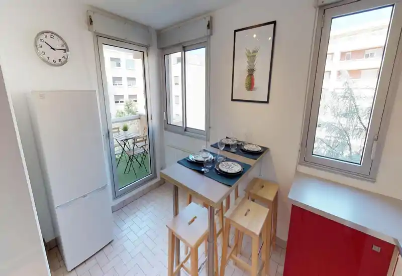 Cucina compatta e luminosa con frigorifero alto, bancone rialzato apparecchiato per quattro persone e grandi finestre che si affacciano su un piccolo balcone. Layout moderno e funzionale con luce naturale e vista gradevole.