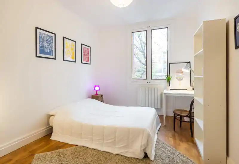 Bright, minimalist bedroom with a neatly made double bed, simple artwork on the wall, a small bedside lamp, and a desk by the window — clean lines and warm wooden floors create a calm atmosphere.