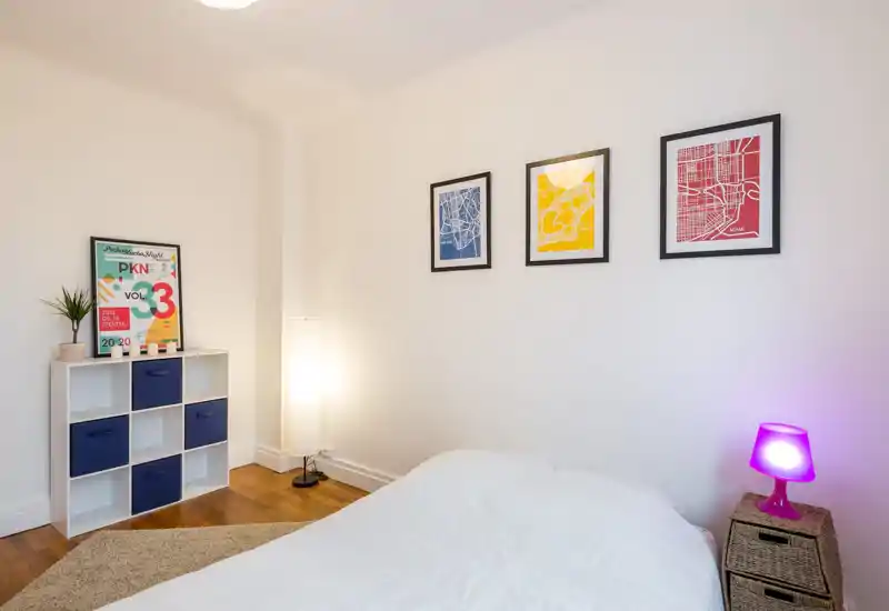 Bright, minimalist bedroom with a neatly made bed, framed artwork on the wall and soft ambient lamps creating a cozy atmosphere.