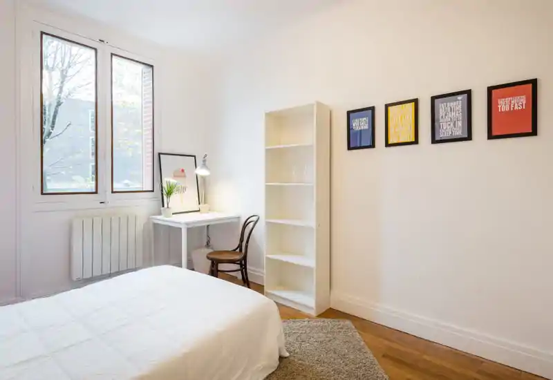 Bright, minimal bedroom with a white bed, natural light from a large window, a small desk and shelving — ideal for a calm, modern rental.
