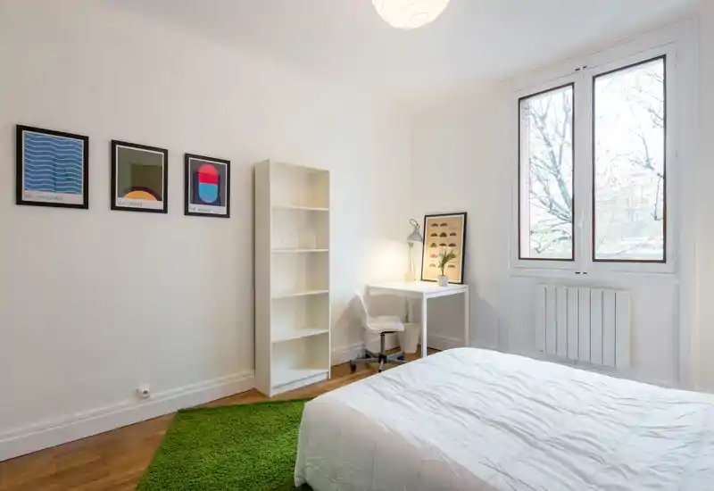 Camera da letto luminosa e minimal con letto bianco, una scrivania vicino alla finestra, scaffale aperto e un tappeto verde che dà un tocco di freschezza.