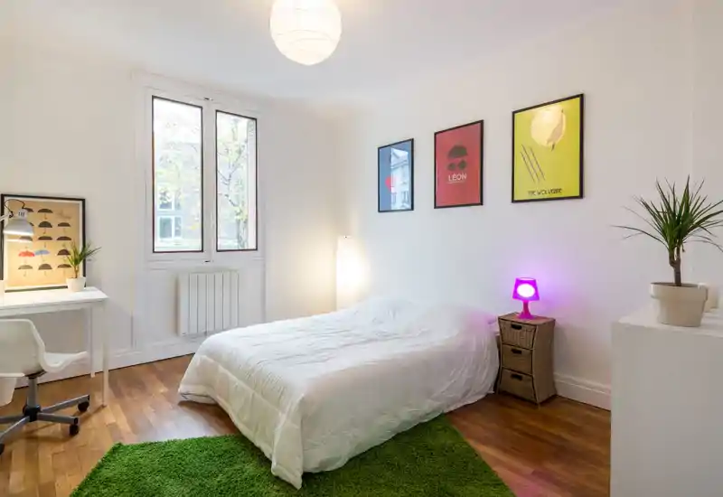 Camera da letto moderna e luminosa con un letto matrimoniale centrale, stampe decorative alle pareti, un tappeto verde e luce naturale da una finestra ampia. Arredata con semplicità: scrivania, piante e lampade soft per un ambiente accogliente.