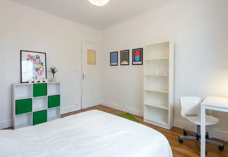 Camera da letto luminosa e minimale con letto ben rifatto in primo piano, scaffale bianco, contenitori verdi e una scrivania piccola: ambiente moderno e arioso.