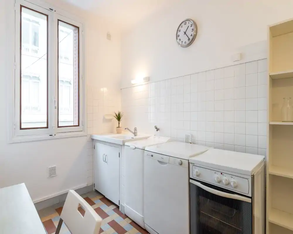 Cuisine compacte et lumineuse avec carreaux blancs, évier, lave-vaisselle et four indépendant ; décoration épurée et petite plante pour une touche accueillante.