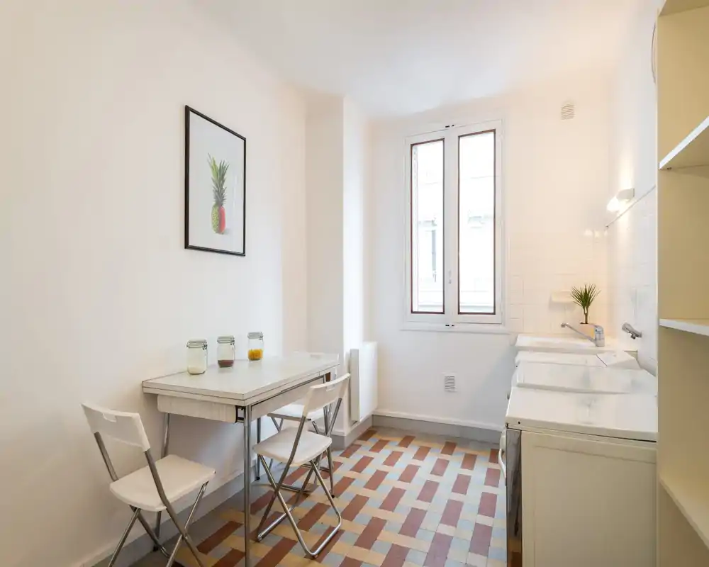 Cuisine lumineuse et minimaliste avec petite table, chaises pliantes et sol carrelé. La lumière naturelle et une décoration sobre donnent un espace propre et fonctionnel.