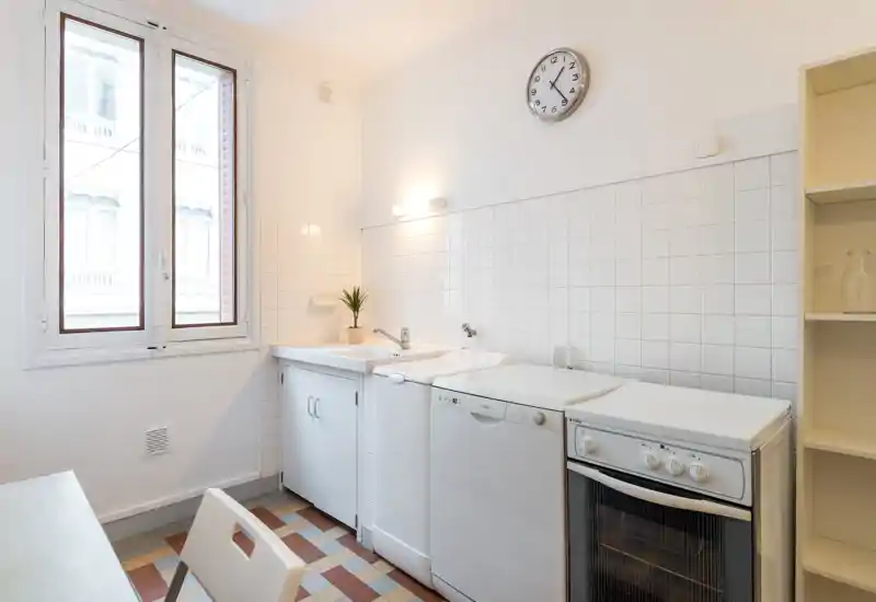 Bright, compact kitchen with white tiles, sink, dishwasher and freestanding oven; clean, minimal styling and a small plant for a welcoming touch.