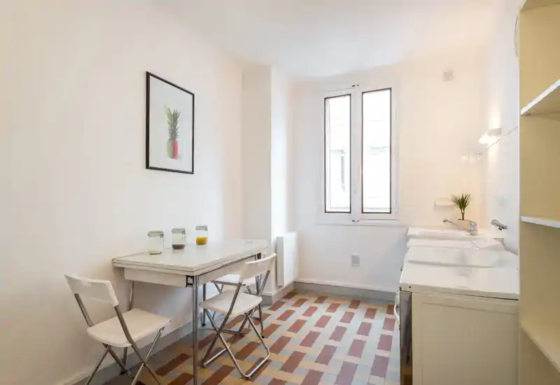 Bright, minimalist kitchen with a small dining table, folding chairs and a tiled floor. Natural light from the window and simple decor create a tidy, functional space.